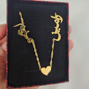 May include: A gold necklace featuring Arabic script pendants and a heart-shaped pendant, presented in a black box with a red patterned edge. This necklace is likely a personalised piece of jewellery.