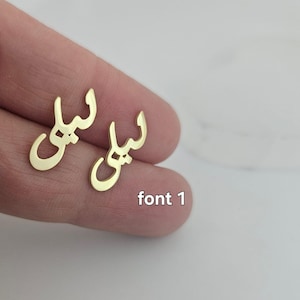 May include: A pair of gold-colored earrings in the shape of the Arabic word "Leila" in font 1. The earrings are displayed on a person's finger against a white background. The text "font 1" is also visible.