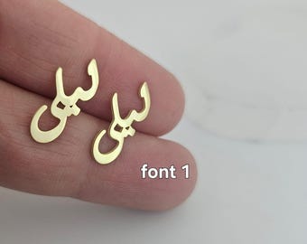 May include: A pair of gold-colored earrings in the shape of the Arabic word "Leila" in font 1. The earrings are displayed on a person's finger against a white background. The text "font 1" is also visible.