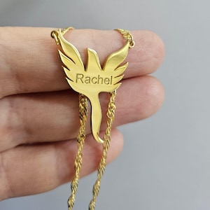 May include: A gold-colored necklace featuring a pendant shaped like a stylized bird with wings spread wide. The name Rachel is engraved in a clear, sans-serif font across the center of the bird's body. The pendant is attached to a twisted chain.