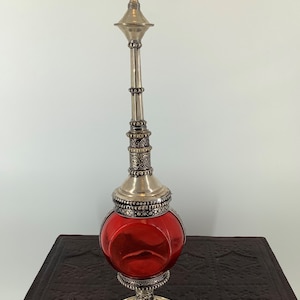 May include: A silver and red glass perfume bottle with intricate detailing. The bottle has a round body and a tall, slender neck with a decorative top.