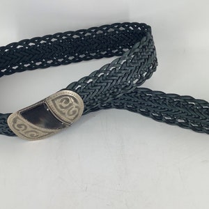 Peut inclure: Une ceinture en cuir tressé noir avec une boucle argentée ornée d'un motif. La boucle a un centre brun foncé.
