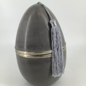 Può includere: Una scatola di ceramica grigia a forma di uovo con una fascia argentata attorno al centro e un fiocco grigio in cima.