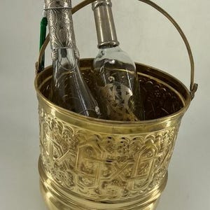 Puede incluir: Un cubo de vino de metal dorado con un diseño en relieve ornamentado. Dentro hay dos botellas de vidrio, una con una tapa plateada decorativa y una borla verde. El cubo tiene un asa y está sobre una superficie blanca.