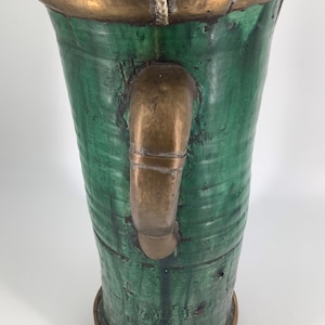 Puede incluir: Un jarrón alto de cerámica verde con un borde y un asa de latón. El jarrón tiene una superficie texturizada y una abertura ligeramente acampanada.
