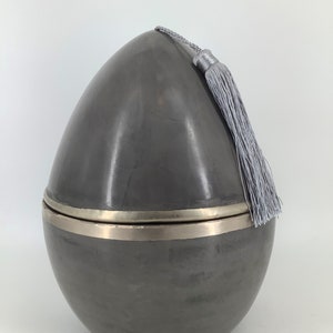 Può includere: Un contenitore in ceramica grigio a forma di uovo con una fascia argentata attorno alla parte superiore e un fiocco grigio. Il contenitore ha un coperchio.