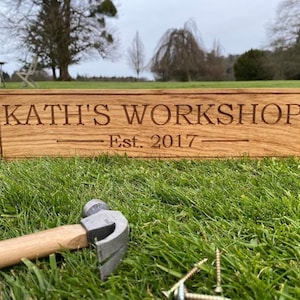 May include: A wooden sign reading "KATH'S WORKSHOP Est. 2017" lies on grass, with a hammer and screws nearby. The sign is rectangular, made of light brown wood, and features engraved text. The scene suggests a workshop setting.
