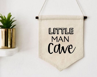 May include: A white canvas banner with the text "LITTLE MAN cave" in black lettering. The banner is hanging from a white wall with a wooden dowel rod at the top.