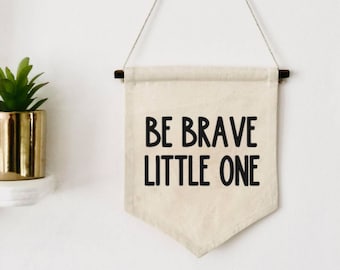 May include: A white canvas banner with black text that reads "Be Brave Little One". The banner is hanging from a white wall with a wooden dowel rod at the top and bottom.