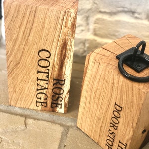 K&ouml;nnte beinhalten: Zwei Holz-T&uuml;rstopper mit Metallringen. Der T&uuml;rstopper links ist mit "ROSE COTTAGE" graviert. Der T&uuml;rstopper rechts ist mit "THE DOOR STOP" graviert.