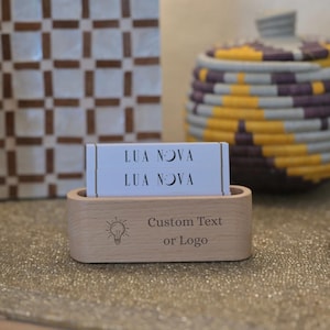 May include: A light wooden business card holder with a lightbulb graphic and the words "Custom Text or Logo". The holder contains white cards with the text "LUA NOVA" and a crescent moon symbol. A decorative basket is in the background.
