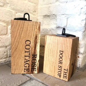 K&ouml;nnte beinhalten: Zwei Holz-T&uuml;rstopper mit schwarzen Metallringen. Der T&uuml;rstopper links ist mit "ROSE COTTAGE" graviert. Der T&uuml;rstopper rechts ist mit "THE DOOR STOP" graviert.