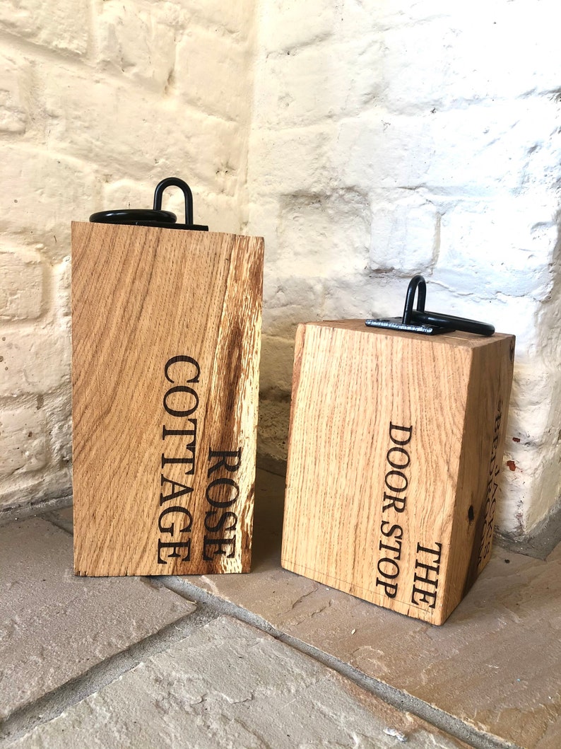 K&ouml;nnte beinhalten: Zwei Holz-T&uuml;rstopper mit schwarzen Metallringen. Der T&uuml;rstopper links ist mit "ROSE COTTAGE" graviert. Der T&uuml;rstopper rechts ist mit "THE DOOR STOP" graviert.