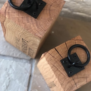 K&ouml;nnte beinhalten: Zwei Holzt&uuml;rstopper mit schwarzen Metallringen. Die T&uuml;rstopper sind aus Naturholz mit rustikalem Finish. Jeder T&uuml;rstopper hat einen schwarzen Metallring, der an einer quadratischen Metallplatte befestigt ist.