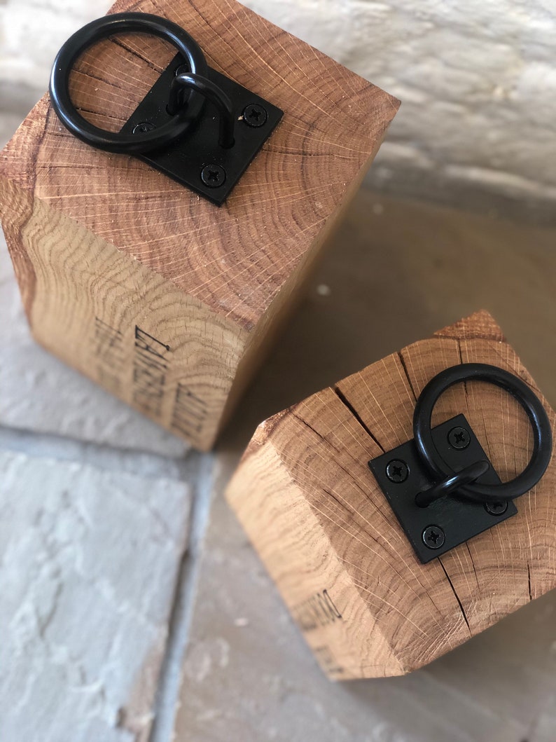 K&ouml;nnte beinhalten: Zwei Holzt&uuml;rstopper mit schwarzen Metallringen. Die T&uuml;rstopper sind aus Naturholz mit rustikalem Finish. Jeder T&uuml;rstopper hat einen schwarzen Metallring, der an einer quadratischen Metallplatte befestigt ist.