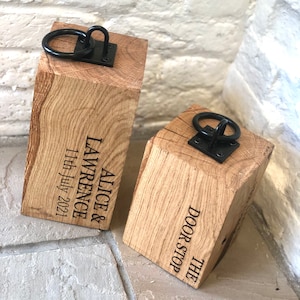 K&ouml;nnte beinhalten: Zwei Holz-T&uuml;rstopper mit schwarzen Metallringen. Der gr&ouml;&szlig;ere T&uuml;rstopper ist mit "Alice & Lawrence 11th July 2021" graviert. Der kleinere T&uuml;rstopper ist mit "The Door Stop" graviert.