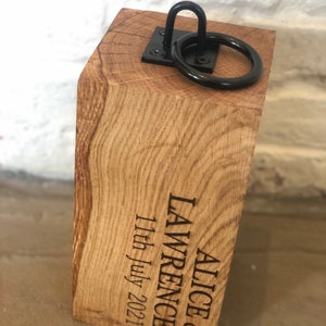 K&ouml;nnte beinhalten: Ein Holz-T&uuml;rstopper mit einem schwarzen Metallring und dem Text "Alice & Lawrence 11th July 2021" auf der Seite eingraviert.