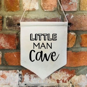 May include: A white canvas banner with black text that reads "LITTLE MAN cave" hanging on a brick wall.