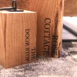 K&ouml;nnte beinhalten: Zwei Holzt&uuml;rstopper mit schwarzen Metallbeschl&auml;gen. Der T&uuml;rstopper links tr&auml;gt die Aufschrift "The Door Stop" und der T&uuml;rstopper rechts die Aufschrift "Rose Cottage".