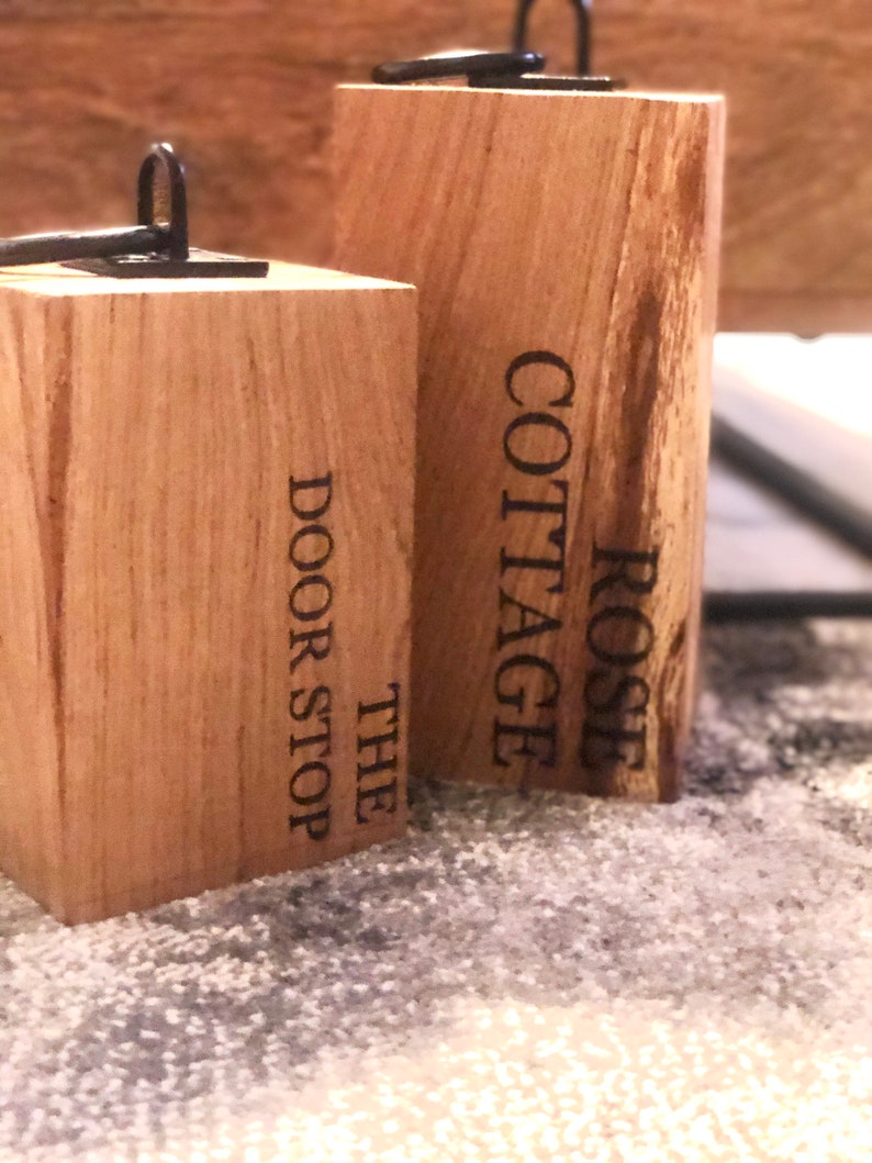 K&ouml;nnte beinhalten: Zwei Holzt&uuml;rstopper mit schwarzen Metallbeschl&auml;gen. Der T&uuml;rstopper links tr&auml;gt die Aufschrift "The Door Stop" und der T&uuml;rstopper rechts die Aufschrift "Rose Cottage".