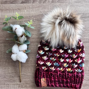 May include: A burgundy knit beanie with a large faux fur pom-pom. The hat features a textured pattern with colorful accents. A small leather tag is attached to the brim. A sprig of cotton and eucalyptus is to the left.