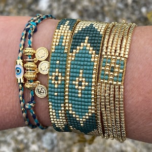 Pode incluir: Uma coleção de pulseiras de contas em tons de verde-azulado e dourado. Uma pulseira apresenta um design tecido, enquanto outras são feitas de pequenas contas douradas. Uma pulseira trançada com pingentes dourados e um pingente de olho grego também está incluída.