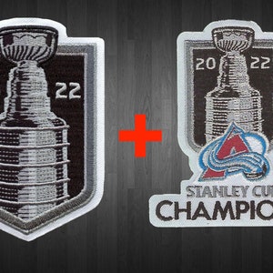 May include: Two embroidered patches featuring the Stanley Cup trophy and the Colorado Avalanche logo. The patches are white with black and silver embroidery. The Stanley Cup patch has the years 20 and 22 embroidered on either side of the trophy. The Avalanche patch has the team's logo and the words "Stanley Cup Champions" embroidered below.