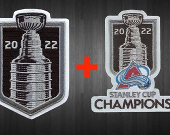 May include: Two embroidered patches featuring the Stanley Cup trophy and the Colorado Avalanche logo. The patches are white with black and silver embroidery. The Stanley Cup patch has the years 20 and 22 embroidered on either side of the trophy. The Avalanche patch has the team's logo and the words "Stanley Cup Champions" embroidered below.