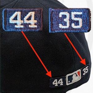 May include: A black baseball cap with a red brim. The cap has two patches on the front, one with the number "44" and the other with the number "35". On the back of the cap, there is a patch with the MLB logo and the numbers "44" and "35".