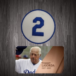 May include: A circular patch with the number 2 in blue on a white background. Below is a photo of Tommy Lasorda, with text that reads "TOMMY LASORDA 1927-2021". The image is set against a wood grain background.