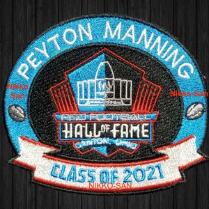 May include: A blue and black embroidered patch with the text "Peyton Manning" in a white circle around the outside. The center of the patch features a black and white image of a football stadium with the text "Pro Football Hall of Fame" and "Class of 2021" in a white banner.