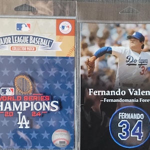 May include: A blue and white World Series Champions patch with a baseball and a stadium silhouette. The patch is on a blue background with white stars. The text "Fernando Valenzuela - Fernandomania Forever" is on a black background with a blue and white baseball patch with the number 34.