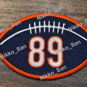 May include: An oval-shaped embroidered patch featuring a dark blue background with a bright orange border. The patch displays the number 89 in white with an orange outline, and a white stitched graphic representing football laces.