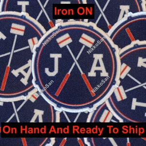 May include: Iron-on patch with the letters "JA" in white, crossed lacrosse sticks in red and white, and a blue background. The patch is surrounded by other patches with the same design.
