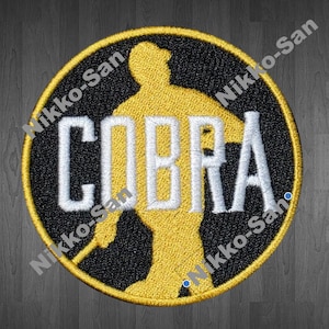 May include: Embroidered patch with a black background and a yellow border. The patch features a yellow silhouette of a person in a karate stance, with the word "COBRA" in white block letters. The patch is circular.