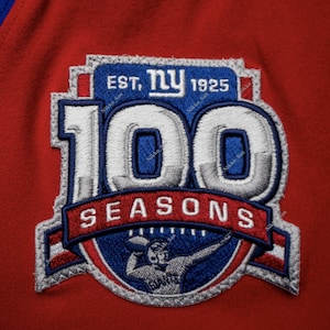 May include: A blue and white embroidered patch with the New York Giants logo and the text "EST. NY 1925 100 SEASONS". The patch features a football player in a blue uniform holding a football.