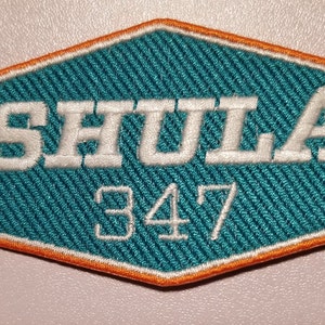 May include: A teal blue and orange embroidered patch with the text "SHULA 347" in white.