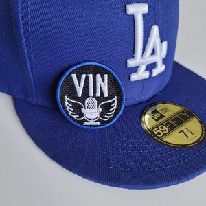 May include: A blue baseball cap with a white "LA" logo on the front. A black and white patch with the word "VIN" and a microphone with wings is attached to the cap.