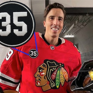 May include: A hockey player wearing a red jersey with a black and white number 35 on the front. The jersey has a black and white number 35 on the shoulder and a black and white logo with a red, white, and black Indian head on the front. The player is holding a hockey stick.