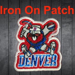 May include: Iron-on patch featuring a cartoon mascot holding a baseball bat and a basketball. The mascot is wearing a blue and red uniform with a white hat and has a red beard. The text "DENVER" is written in blue and white on a red background.