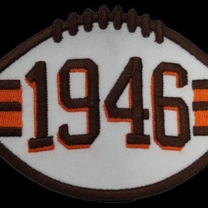 May include: A white football shaped patch with brown and orange stripes and the number 1946 in brown.