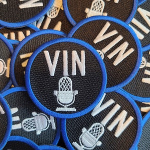 May include: A close-up of several black and blue circular patches with white embroidery. The patches feature the word "VIN" and a microphone graphic.