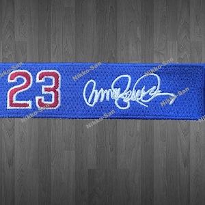 May include: A blue rectangular patch with the number "23" in red and white, and a white signature. The patch is embroidered with a textured appearance. The background is a dark wood grain.