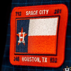 May include: A red, white, and blue embroidered patch with the text "SPACE CITY 201" at the top and "HOUSTON, TX" at the bottom. The patch features a stylized star with the letter "H" inside, representing the Houston Astros baseball team.