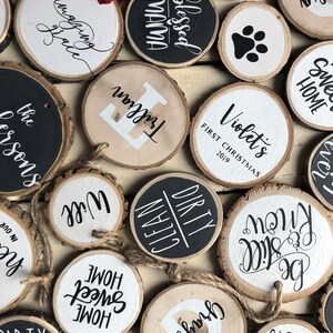 May include: A collection of round wood slices with various painted designs and text. Some of the designs include "Sweet Home", "Blessed Mama", "Clean Dirty", "Will", "Trillian", "Violet's First Christmas 2019", and "Be Still Know".