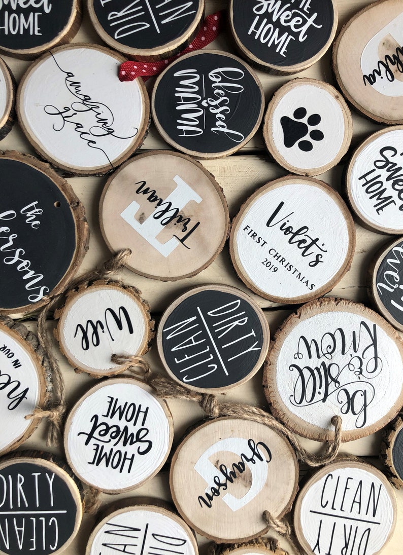 May include: A collection of round wood slices with various painted designs and text. Some of the designs include "Sweet Home", "Blessed Mama", "Clean Dirty", "Will", "Trillian", "Violet's First Christmas 2019", and "Be Still Know".