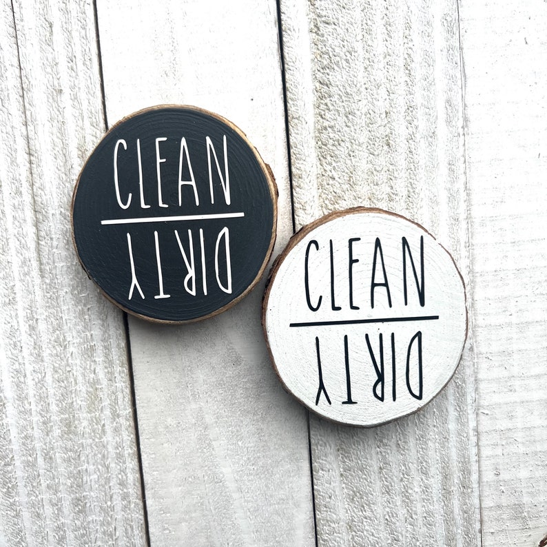 May include: Two round wooden signs with the words "Clean" and "Dirty" written on them in black and white. The signs are on a white wooden background.