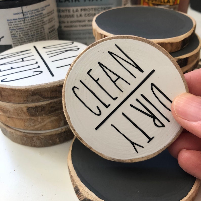 May include: A set of round wooden signs with a white background and black lettering. The signs say "Clean" and "Dirty" with a line separating the words. The signs are perfect for organizing and labeling.