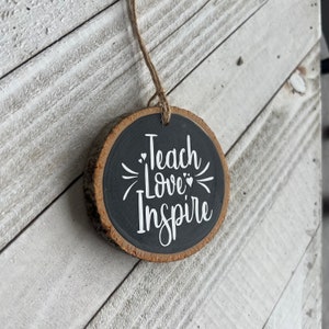 May include: A round wooden ornament with a black painted surface and the words "Teach Love Inspire" written in white. The ornament is hanging from a string.