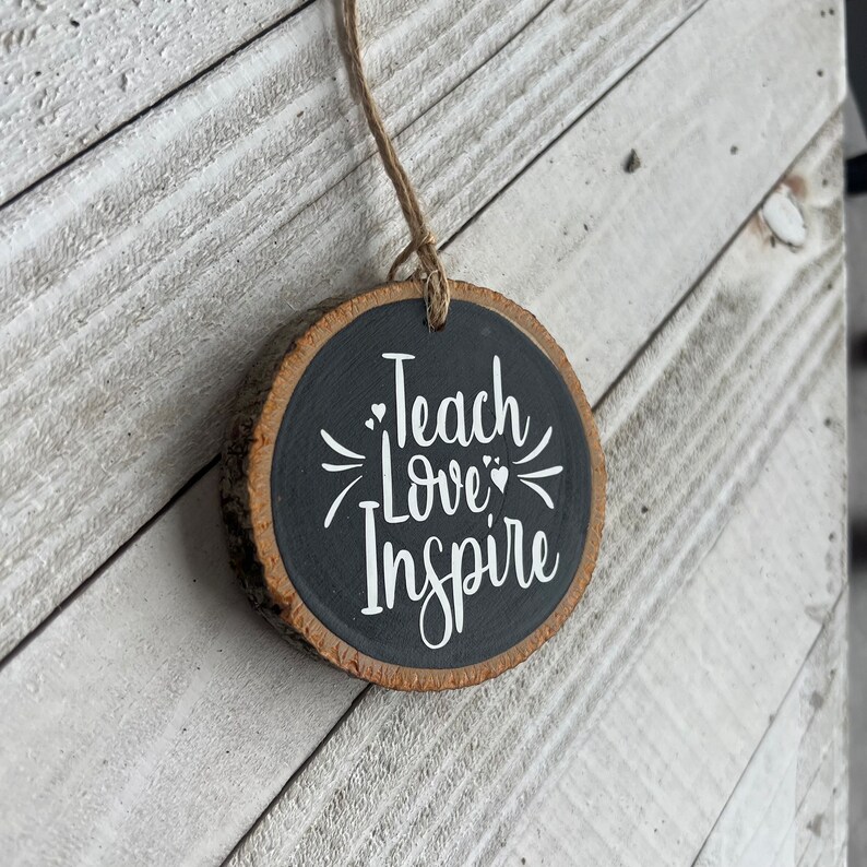 May include: A round wooden ornament with a black painted surface and the words "Teach Love Inspire" written in white. The ornament is hanging from a string.